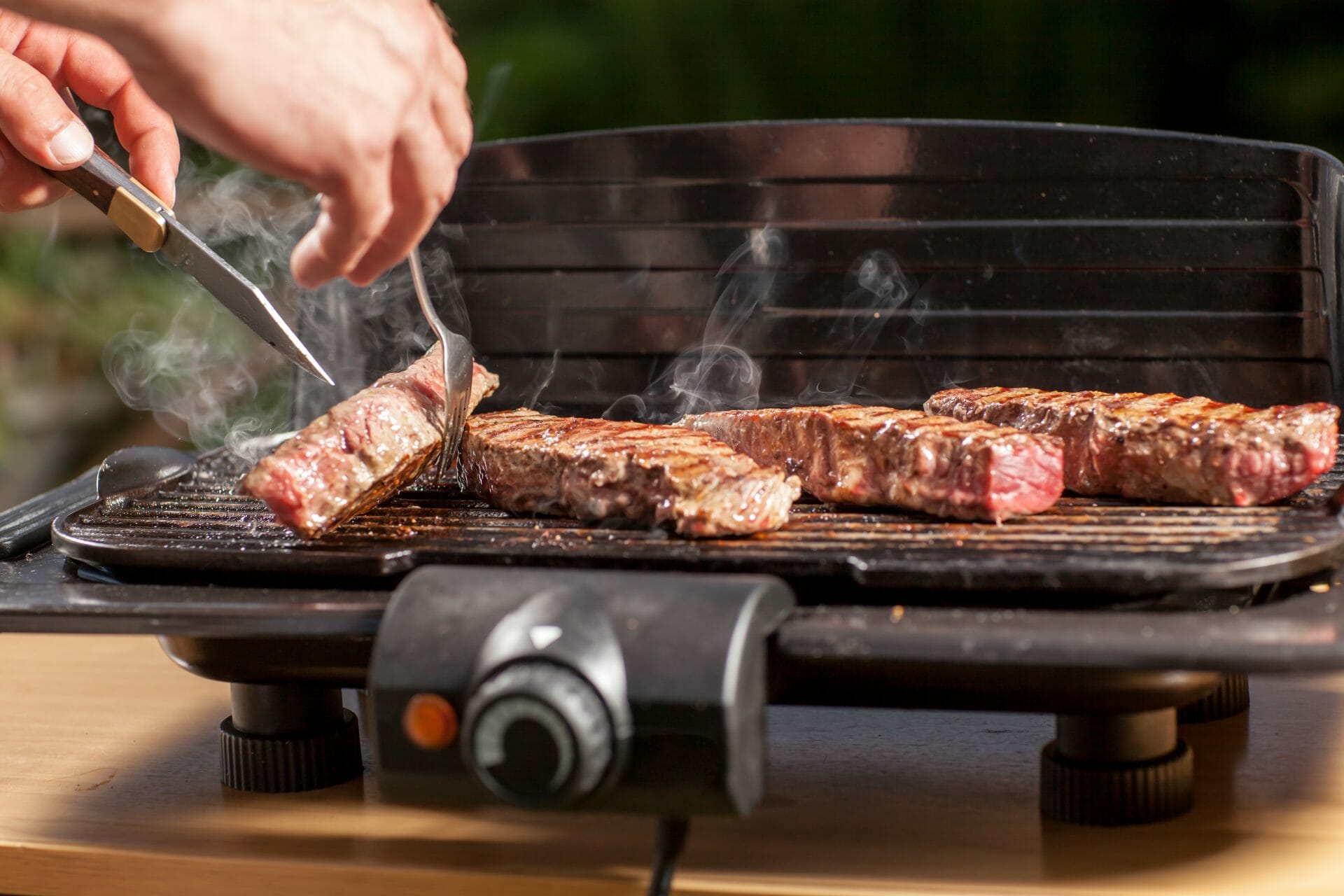 electric grill on counter with red meat on it and person's hands flipping one steak